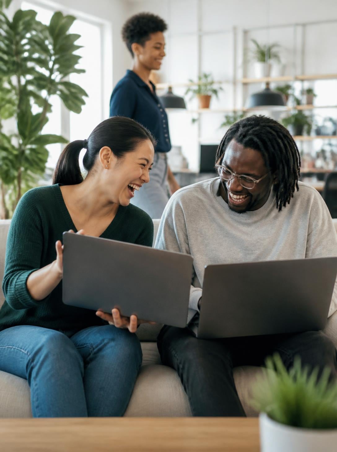 Zwei Kolleg:innen sitzen lachend mit ihren Laptops auf einem Sofa, im Hintergrund eine dritte Person